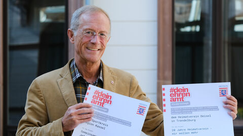 Staatsminister Wintermeyer hält zwei Förderbescheide "#deinehrenamt" für Projekte im Landkreis Kassel in die Kamera und lächelt.