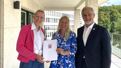 Sportministerin Stolz steht zwischen einer Frau und einem Mann. Sie übergibt der Dame links von sich einen Förderbescheid und alle lächeln in die Kamera.