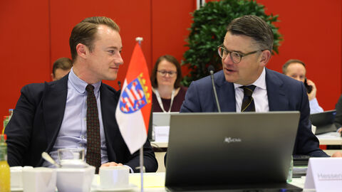 Zwei Männer sitzen in Anzügen nebeneinander und schauen sich an. Vor ihnen steht auf dem Tisch ein Laptop.